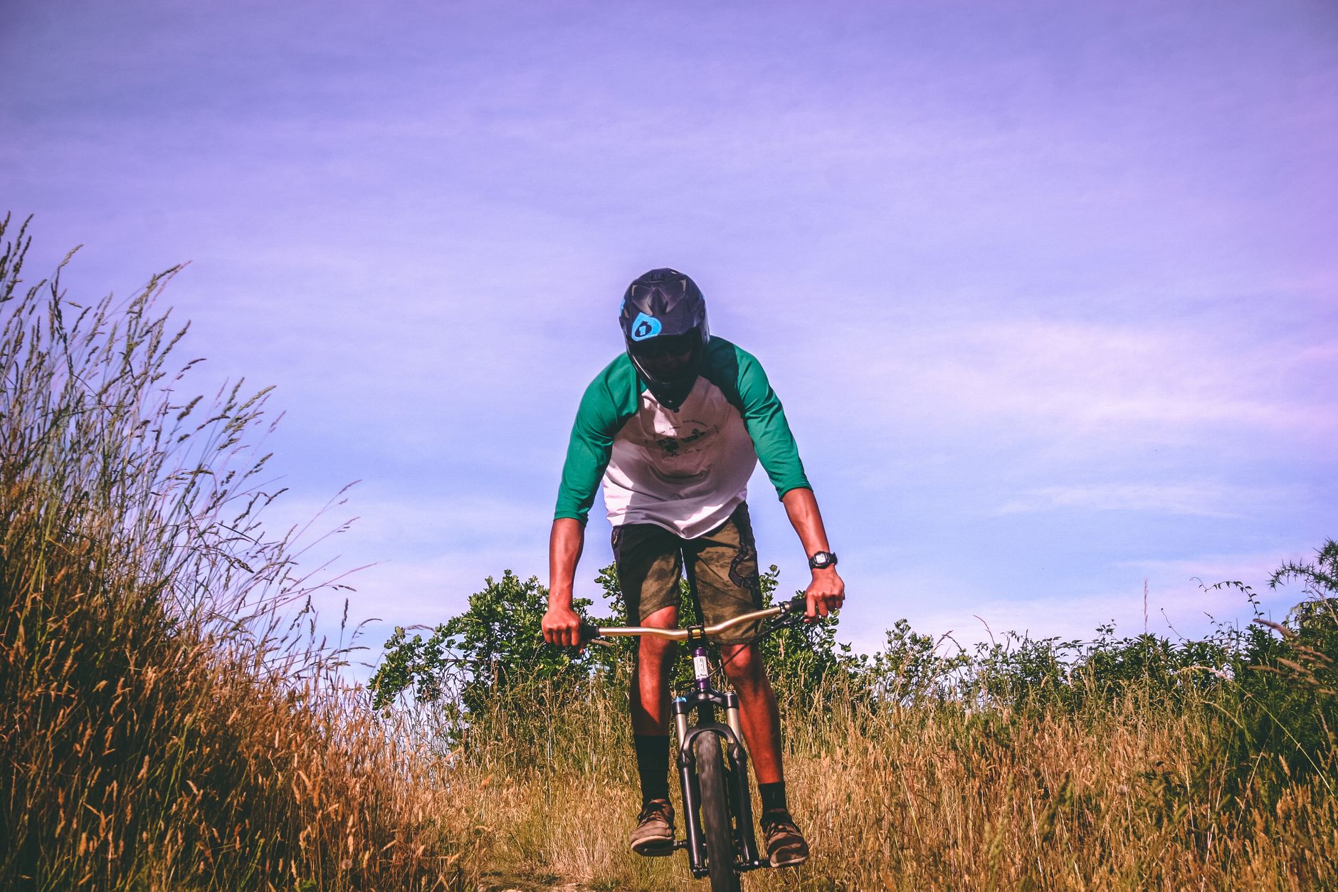 Cyclist riding through dry grass field wearing green jersey, brown vest, and blue helmet against purple sky
