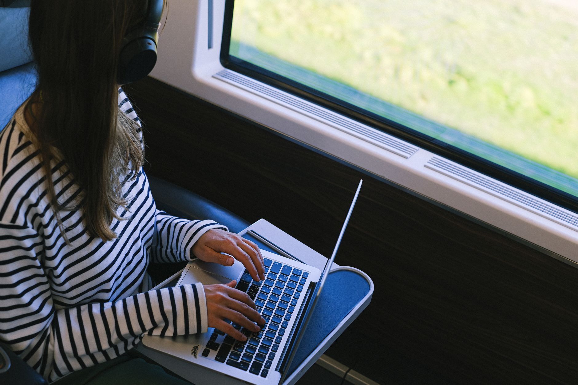 Person in striped shirt typing on laptop by a sunny window on public transport