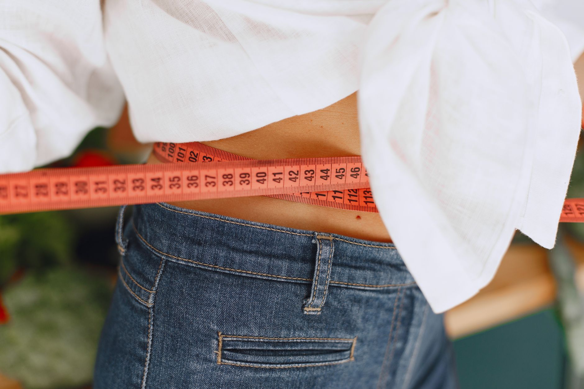 Person in white crop top and jeans measuring waist with orange tape measure