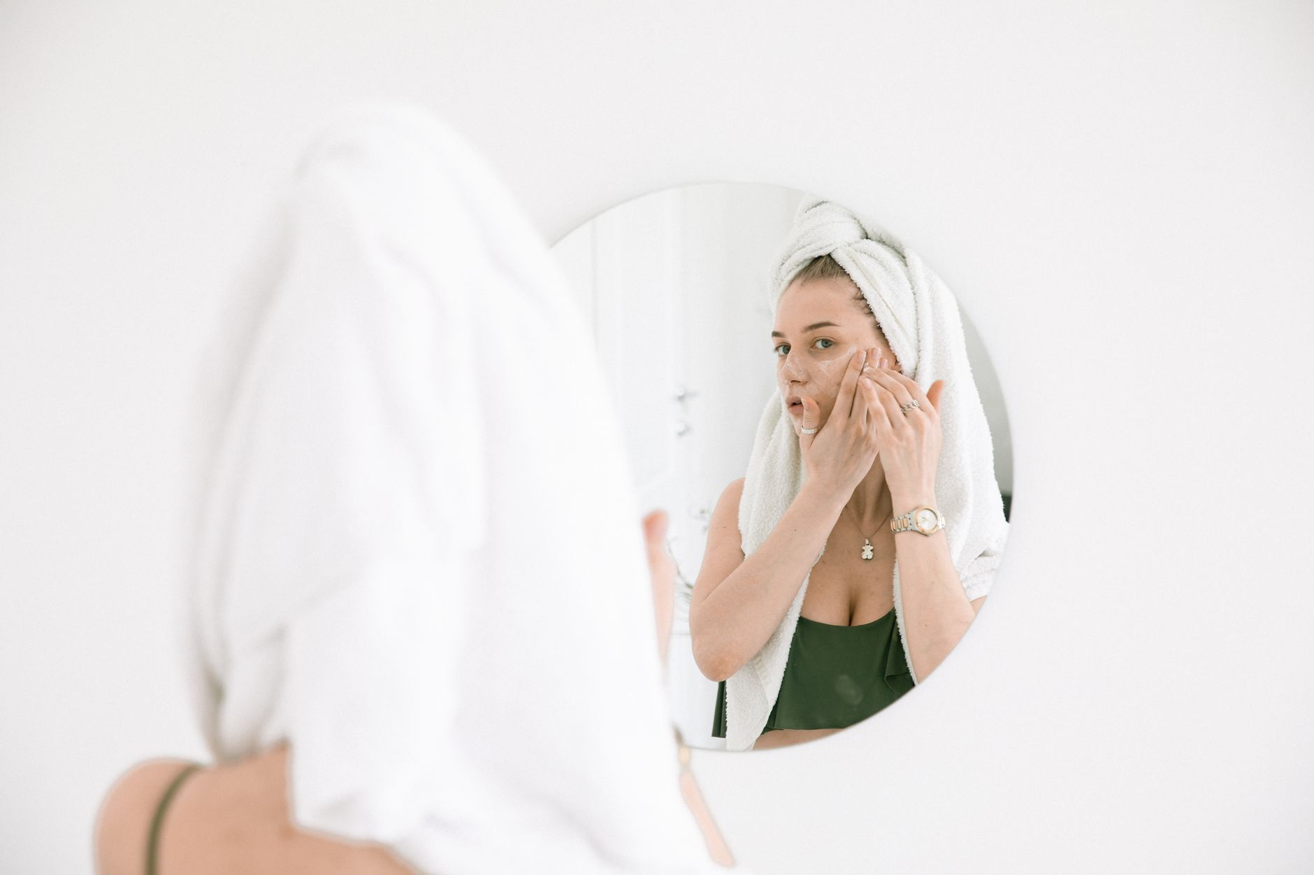 Woman with towel wrapped around head examining skin in round mirror