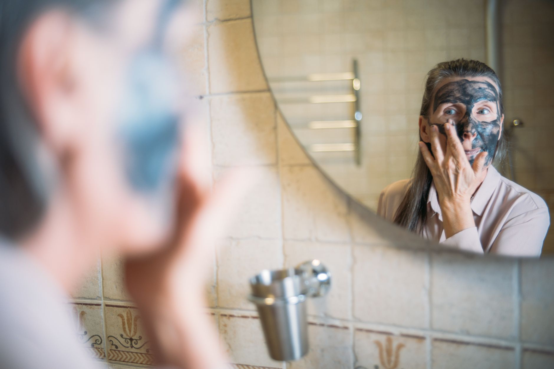 Woman applying dark face mask in bathroom mirror with surprised expression