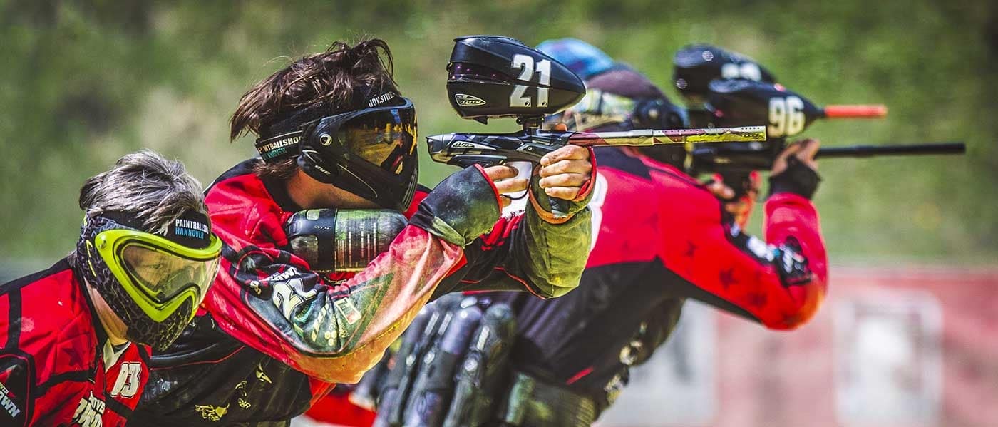 Paintball-Spieler in roten und schwarzen Uniformen mit Masken und Markierern bei Tageslichteinsatz.